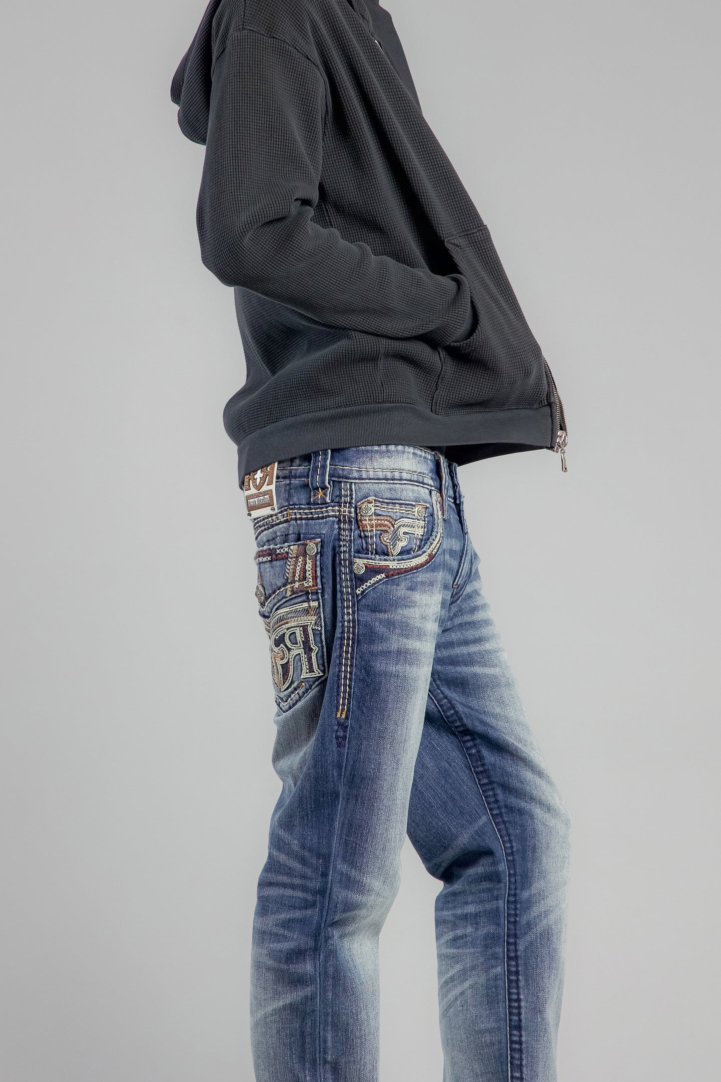 Person wearing a black hoodie and blue jeans with decorative stitching on a gray background