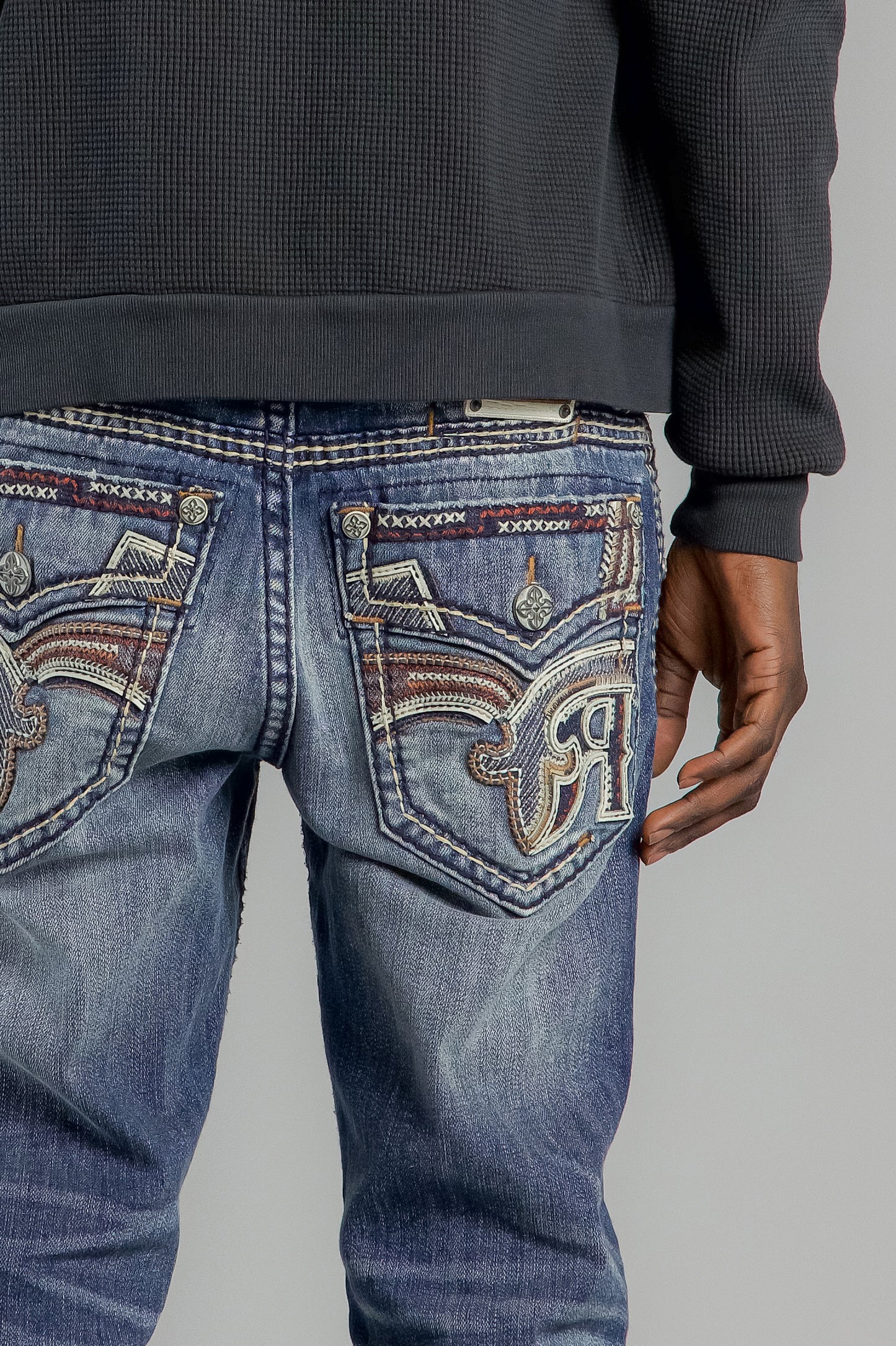 Close-up of blue jeans with intricate patterns on the back pocket against a gray background.